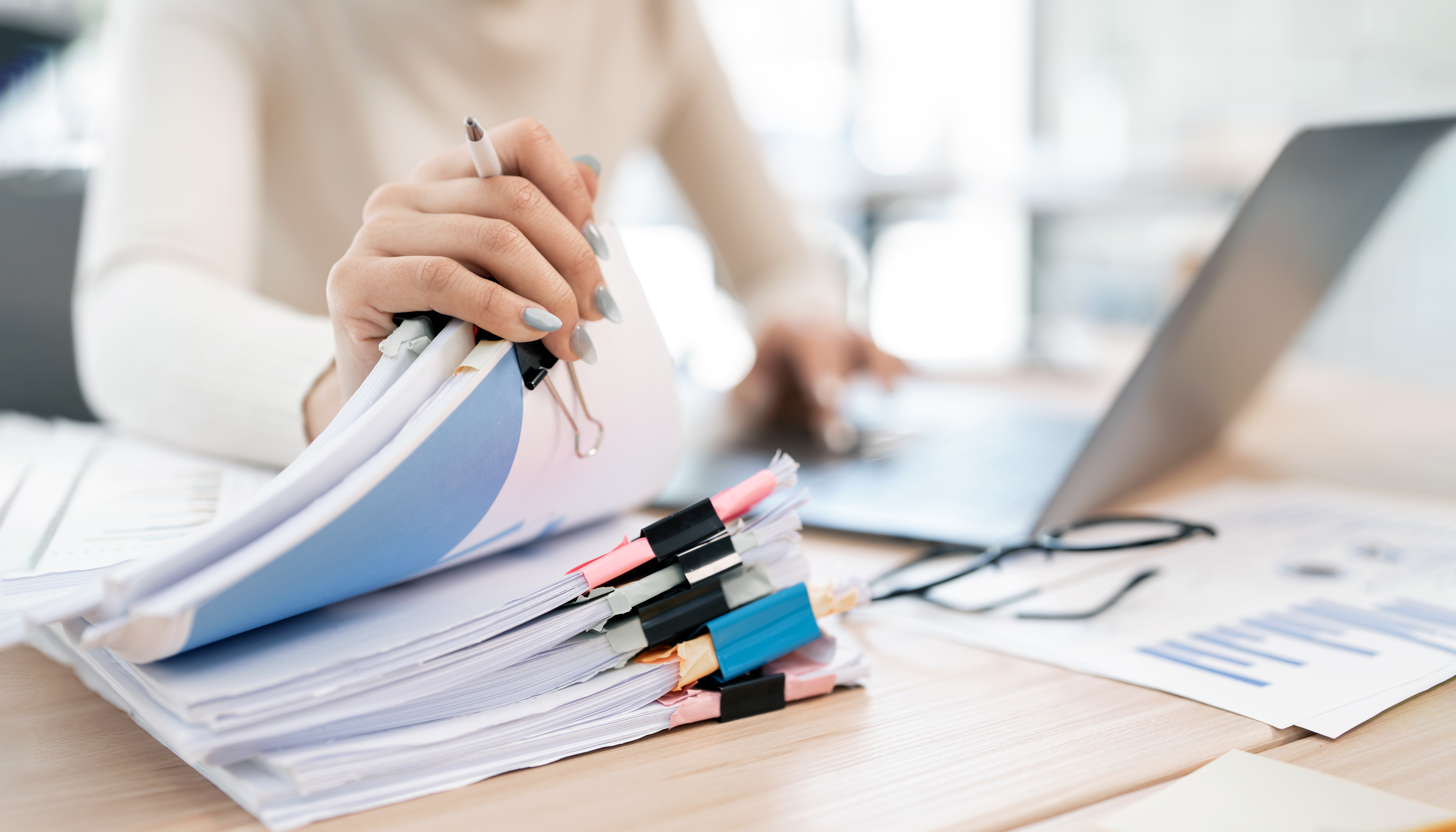 A woman is seen multitasking, holding a stack of organized documents with a pen while working on a laptop.